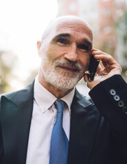 Bearded senior businessman in formal outfit talking on cellphone while walking on street