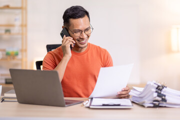 Smiling Asian man writing notes while making phone call and using laptop at home