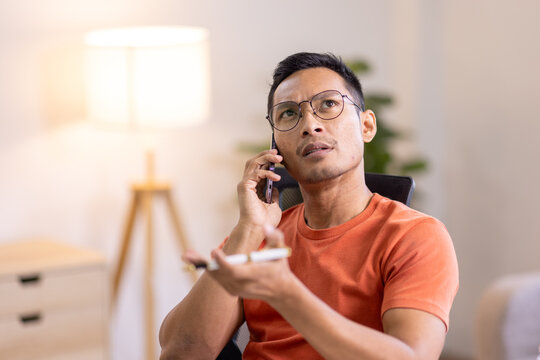 Smiling Asian man writing notes while making phone call and using laptop at home - Powered by Adobe