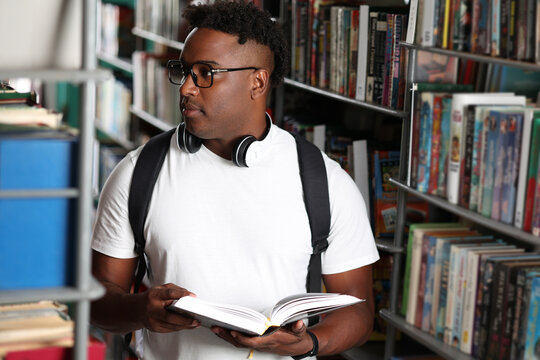Young black man in modern clothes with a backpack is immersed in learning in a library, surrounded by books. Handsome stylish african american man choosing and reading books in modern bookstore