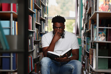 Handsome, stylish young African is reading a book among the bookshelves in the library, adjusting his reading glasses. Cute student with glasses is studying a new book with a serious look - obrazy, fototapety, plakaty