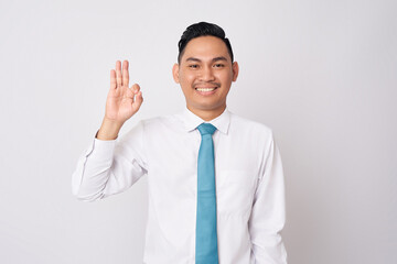 Portrait of a smiling happy young Asian man in formal wear standing confident while showing an ok sign with a finger and looking at camera with smile isolated on white background