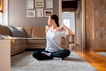 Fototapeta na wymiar Young Caucasian woman wearing sports clothes sits on floor in living room and turns to left side for stretching shoulders. Home yoga and fitness