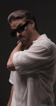 attractive young man in white shirt with sunglasses touching shoulder and moving, crossing arms and posing in a confident way on grey background
