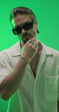 handsome unshaved guy with sunglasses touching chin and thinking, rubbing palms and looking to side, adjusting shirt in front of colorful lights background 