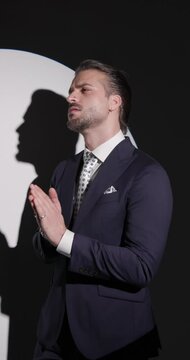 side view video of handsome bearded man looking to side, rubbing palms and fixing suit, touching chin and being thoughtful in front of round spotlight on grey background