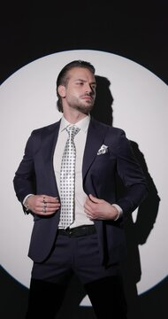 vertical video of confident high class man looking to side, closing suit and smiling, holding hands in pockets and looking down in front of grey background