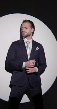 sexy bearded man arranging sleeves and suit while looking to side, unbuttoning suit, touching chin and thinking in front of grey background