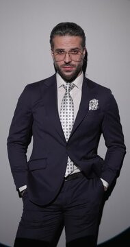 confident bearded man in suit holding hands in pockets, looking to side and posing in a serious way on grey background
