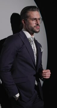 vertical video of handsome bearded businessman holding hand in pocket, adjusting and unbuttoning suit, looking forward and to side on grey background