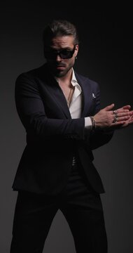vertical video of sexy elegant man with sunglasses holding hand in pocket, rubbing palms, touching shoulder and moving to side on grey background