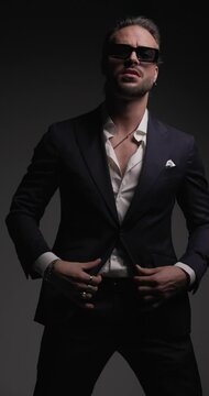 cool unshaved man holding hands in pockets, adjusting suit, and sunglasses, looking to side, pointing fingers forward before leaving on grey background