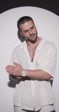 unshaved cool man with glasses with hand in pocket, looking down and side, rubbing palms and moving in a side view pose while leaning on a grey wall