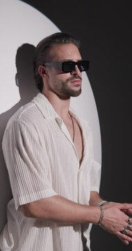 cool young man in white shirt looking forward and adjusting sunglasses, rubbing palms and being confident on grey background