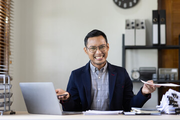 Fototapeta premium Texting to colleague or play games. Confident young asian man in smart casual wear holding smart phone and looking at it while sitting at his working place in office