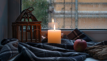 Autumn still life. A burning candle, a wicker cage, a maple leaf and an apple on a warm wool blanket on the windowsill against the background of a window on a rainy day.