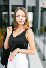 A girl with long blond hair, wearing a black top, black jacket and white shorts, poses against the backdrop of modern architecture. Young woman walking in a park with landscape design