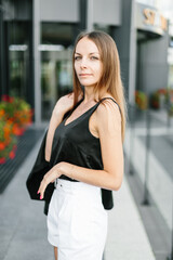Fototapeta premium A girl with long blond hair, wearing a black top, black jacket and white shorts, poses against the backdrop of modern architecture. Young woman walking in a park with landscape design