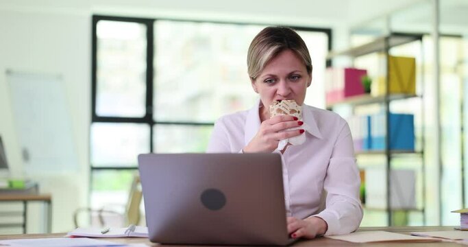 Woman manager eating shawarma at table in front of laptop 4k movie slow motion. Meal break at work concept 