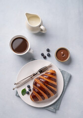Fresh croissant with chocolate and blueberries on a white plate on a light background with a cup of black coffee and milk jug. Delicious homemade breakfast concept.