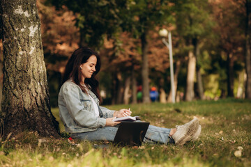 Young businesswoman working on her notebook in park. Freelancer woman sitting on foliage under tree using laptop computer for work. Copy space