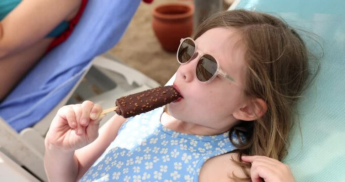 Little girl in sunglasses eating ice cream on sun lounger on beach 4k movie slow motion. Holidays at resort with children concept