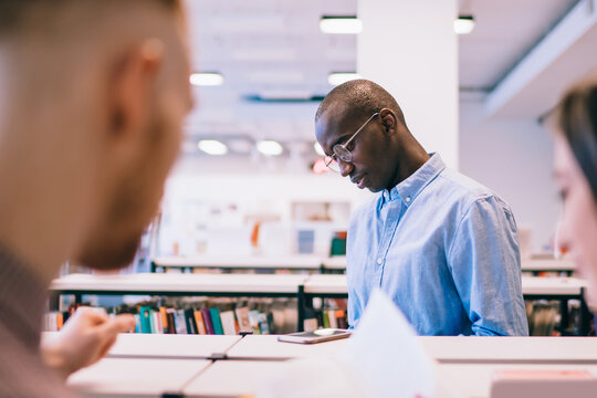 Interested ethnic student reading textbook while standing beside bookshelf in university