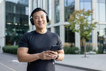 Portrait of a young Asian man standing on a city street wearing headphones and listening to music on the phone. Plays sports, runs, smiles and looks at the camera