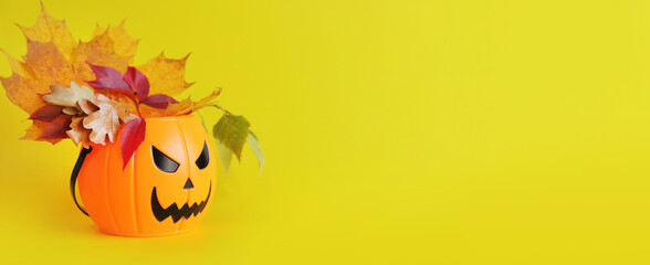 Naklejka na ściany i meble Happy Halloween. Plastic pumpkin bucket with an evil smile on a yellow background, empty space for text. Copy space. Banner for Halloween. A bouquet of autumn leaves in a festive candy bucket