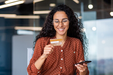 Young successful smiling woman at workplace, using app on phone and bank credit card for online shopping in online store, business woman inside office sitting at table