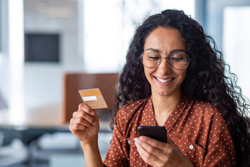 Young successful smiling woman at workplace, using app on phone and bank credit card for online...