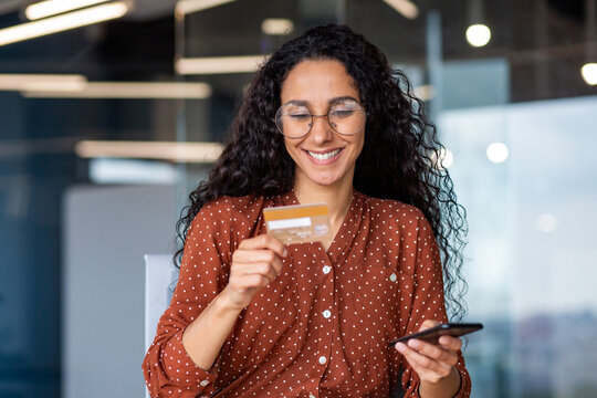 Young successful smiling woman at workplace, using app on phone and bank credit card for online shopping in online store, business woman inside office sitting at table