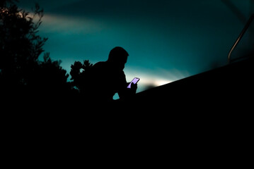 Rear silhouette of a man sitting by the pool at night and looking at his smartphone. Soft focus, silhouette photo