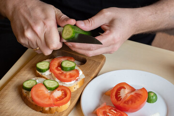 Cooking sandwiches. Man cuts cucumber. Sandwiches with fresh vegetables. Guy is preparing snacks for lunch. Man cuts cucumbers by weight. Quick homemade lunch. Sandwiches on cutting board
