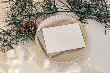 Christmas still life. Blank horizontal greeting card, invitation mockup on scalloped plate in sunlight. Pine cone, pine tree branches. White linen tablecloth, silk ribbon. Winter gift concept. Flatlay