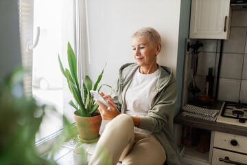 Charming stylish sporty female of 50 sitting on windowsill at kitchen browsing social media on...