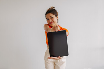 Obraz premium Thai asian woman with bun hair wear orange cropped t-shirt, holding and showing tablet computer black screen, isolated on white background. Smiling and looking at camera.