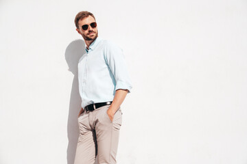 Handsome stylish hipster lambersexual model. Modern man dressed in white shirt and trousers. Fashion male posing in the street in sunglasses near white wall