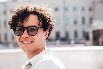 Portrait of young attractive man with curly hair hairstyle. Smiling handsome male in casual stylish clothes posing in the street at sunny day. Cheerful and happy model outdoors. In spectacles, glasses