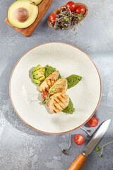 Top view of grilled chicken fillet accompanied by avocado and spinach, elegantly served on a ceramic plate. The grey concrete background and hard shadows add depth to this healthy dish