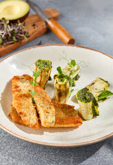 Close-up vertical side view of tempura perch fillet with zucchini rolls on a ceramic plate. The grey concrete background with harsh shadows emphasizes the textures and colors in this gourmet dish