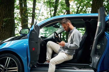 With smartphone in hands. Beautiful young man is sitting in the blue car outdoors