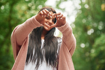 Showing heart gesture by hands. Cheerful beautiful woman is standing outdoors, green leaves at background