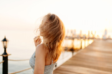 A slender girl with long hair in a gray shiny dress walks along the pier by the sea at sunset. A...