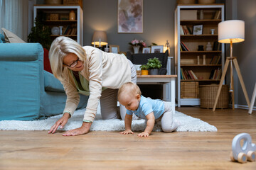 Grandmother of single mother taking care of her young baby, playing on the apartment floor. Senior woman babysitting her grandson or granddaughter while child parent are at work or date night. - obrazy, fototapety, plakaty