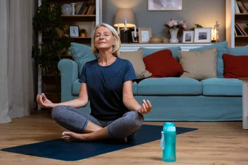 Foto op Canvas Senior woman exercising while sitting in lotus position. Active mature woman doing stretching exercise in living room at home. Fit lady stretching arms and back while sitting on yoga mat. © Srdjan