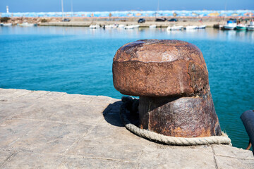 Rusty old steel mooring bollard with sea and fishing boats background.