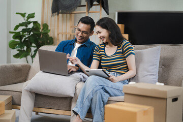 Young couple relaxing sitting on the sofa using the computer laptop around cardboard boxes, very happy moving to a new house.