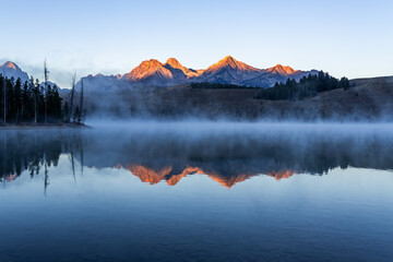 Redfish lake in Idaho in dawn. Calm water covered by mist. Mountains range is reflecting in lake water - Powered by Adobe