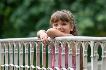 A small, cute, laughing girl of 6 years old with long hair stands on a white bridge in the park and looks at the camera with great curiosity. Close-up portrait